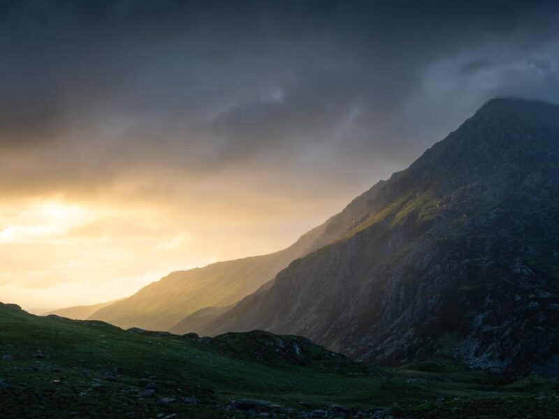 photograph of mountains with a stormy sky above and golden light streaming through on the horizon.